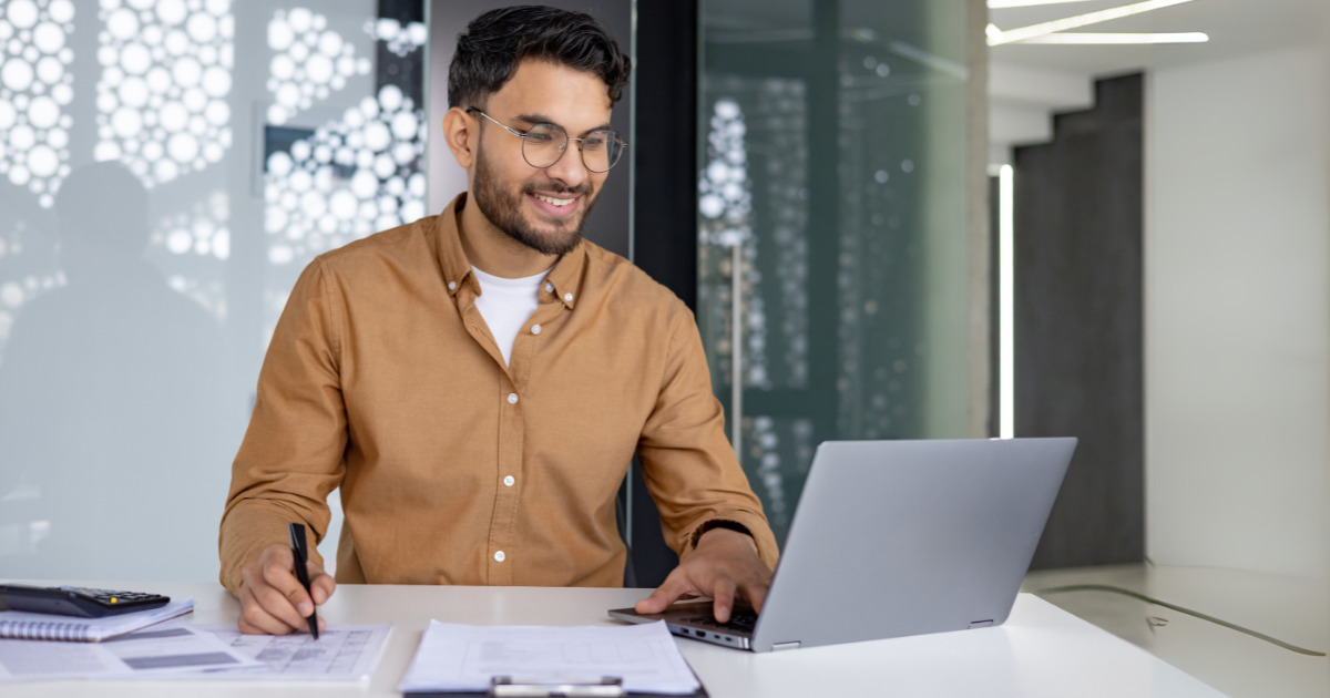 man working on a laptop in the office, sitting at a table writing documents and talking on a video call